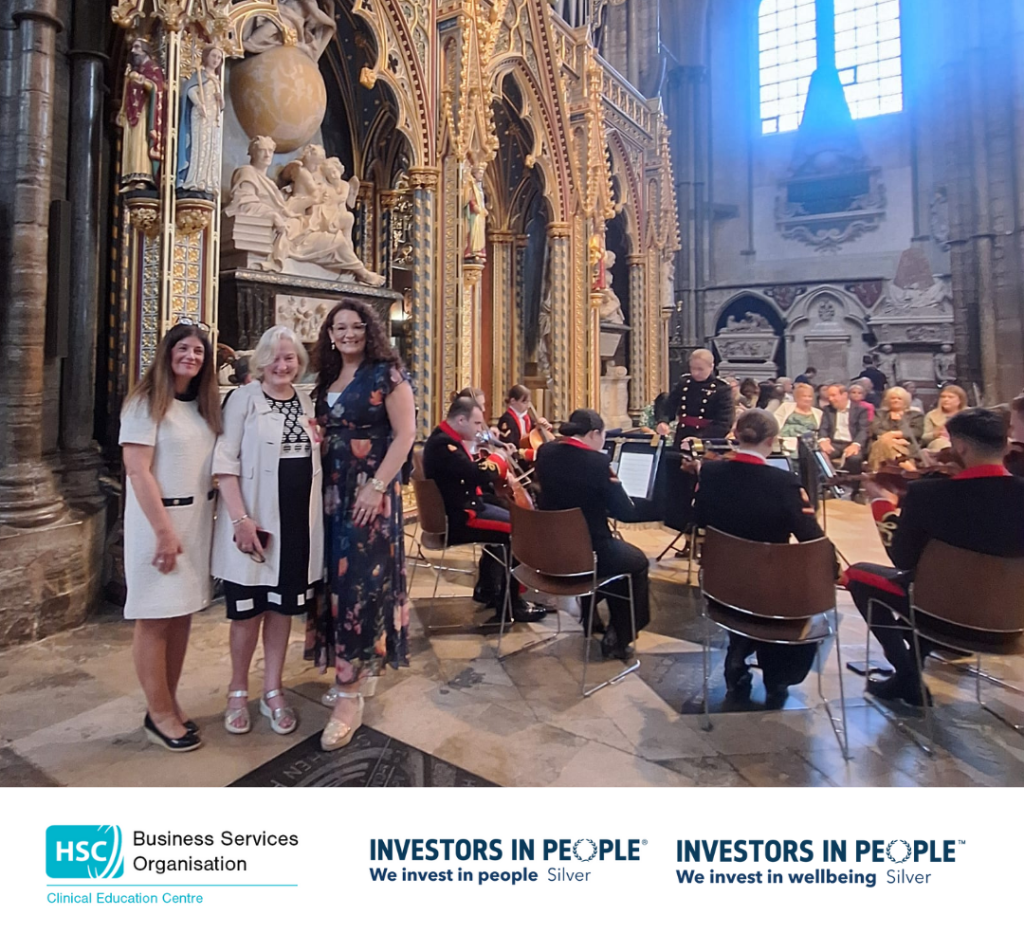 Three females standing in front of ornate architecture with a string orchestra and people seated in the background