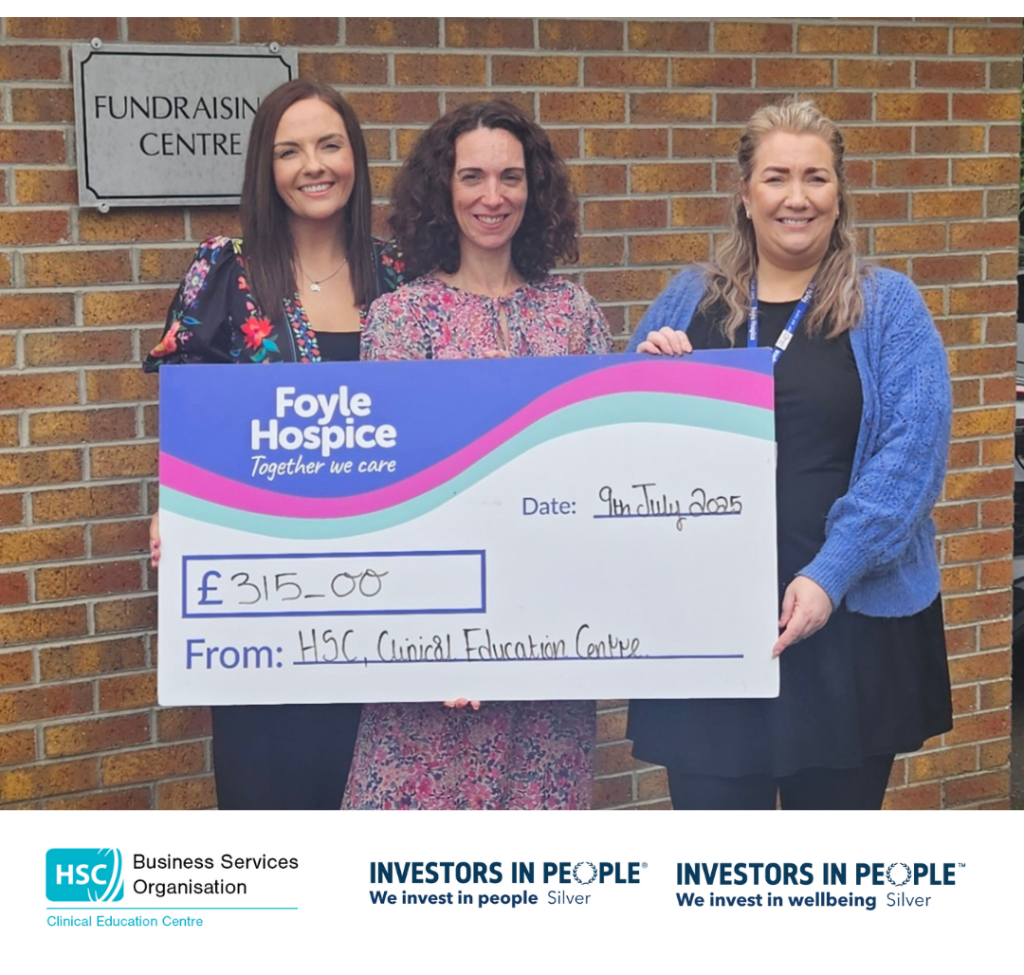Three females standing in front of a brick building holding a large cheque