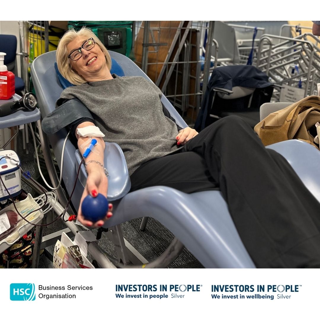 BSO Colleagues leading by example and are reclining on a chair, donating blood. They are holding a blue stress ball and appear relaxed, smiling at the camera. Medical equipment is visible nearby. The bottom of the image features logos for the Business Services Organisation and Investors in People, along with the text "We invest in people Silver" and "We invest in wellbeing Silver.