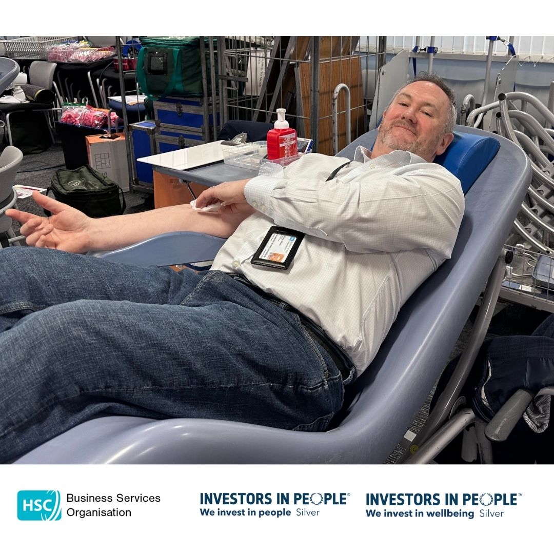 BSO Colleagues leading by example and are reclining on a chair, donating blood. They are holding a blue stress ball and appear relaxed, smiling at the camera. Medical equipment is visible nearby. The bottom of the image features logos for the Business Services Organisation and Investors in People, along with the text "We invest in people Silver" and "We invest in wellbeing Silver.