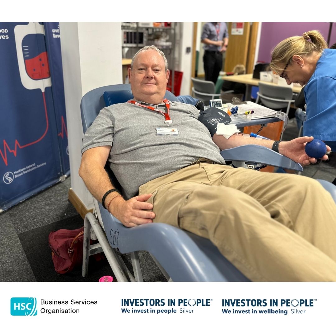 BSO Colleagues leading by example and are reclining on a chair, donating blood. They are holding a blue stress ball and appear relaxed, smiling at the camera. Medical equipment is visible nearby. The bottom of the image features logos for the Business Services Organisation and Investors in People, along with the text "We invest in people Silver" and "We invest in wellbeing Silver.