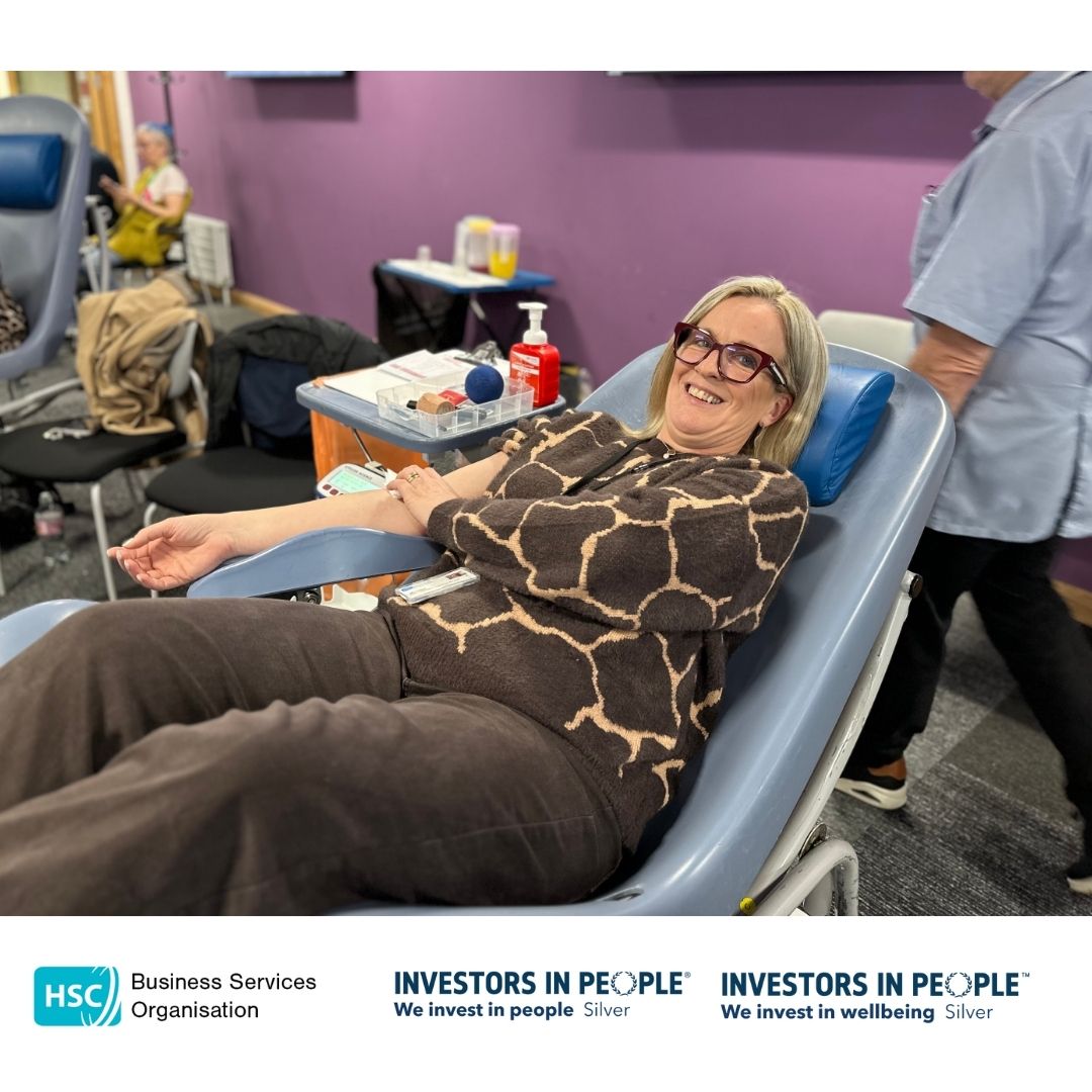 BSO Colleagues leading by example and are reclining on a chair, donating blood. They are holding a blue stress ball and appear relaxed, smiling at the camera. Medical equipment is visible nearby. The bottom of the image features logos for the Business Services Organisation and Investors in People, along with the text "We invest in people Silver" and "We invest in wellbeing Silver.