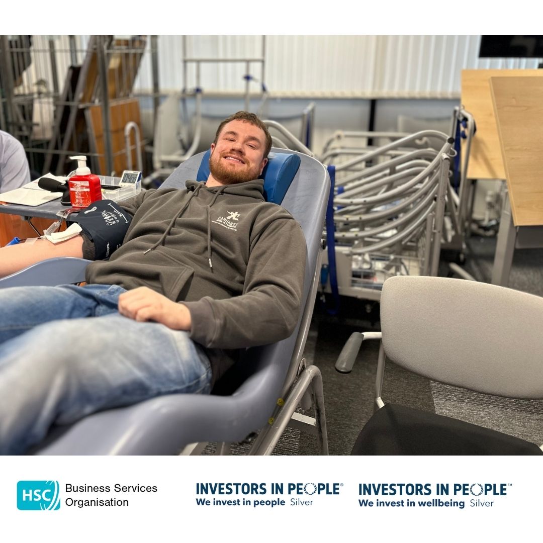 BSO Colleagues leading by example and are reclining on a chair, donating blood. They are holding a blue stress ball and appear relaxed, smiling at the camera. Medical equipment is visible nearby. The bottom of the image features logos for the Business Services Organisation and Investors in People, along with the text "We invest in people Silver" and "We invest in wellbeing Silver.