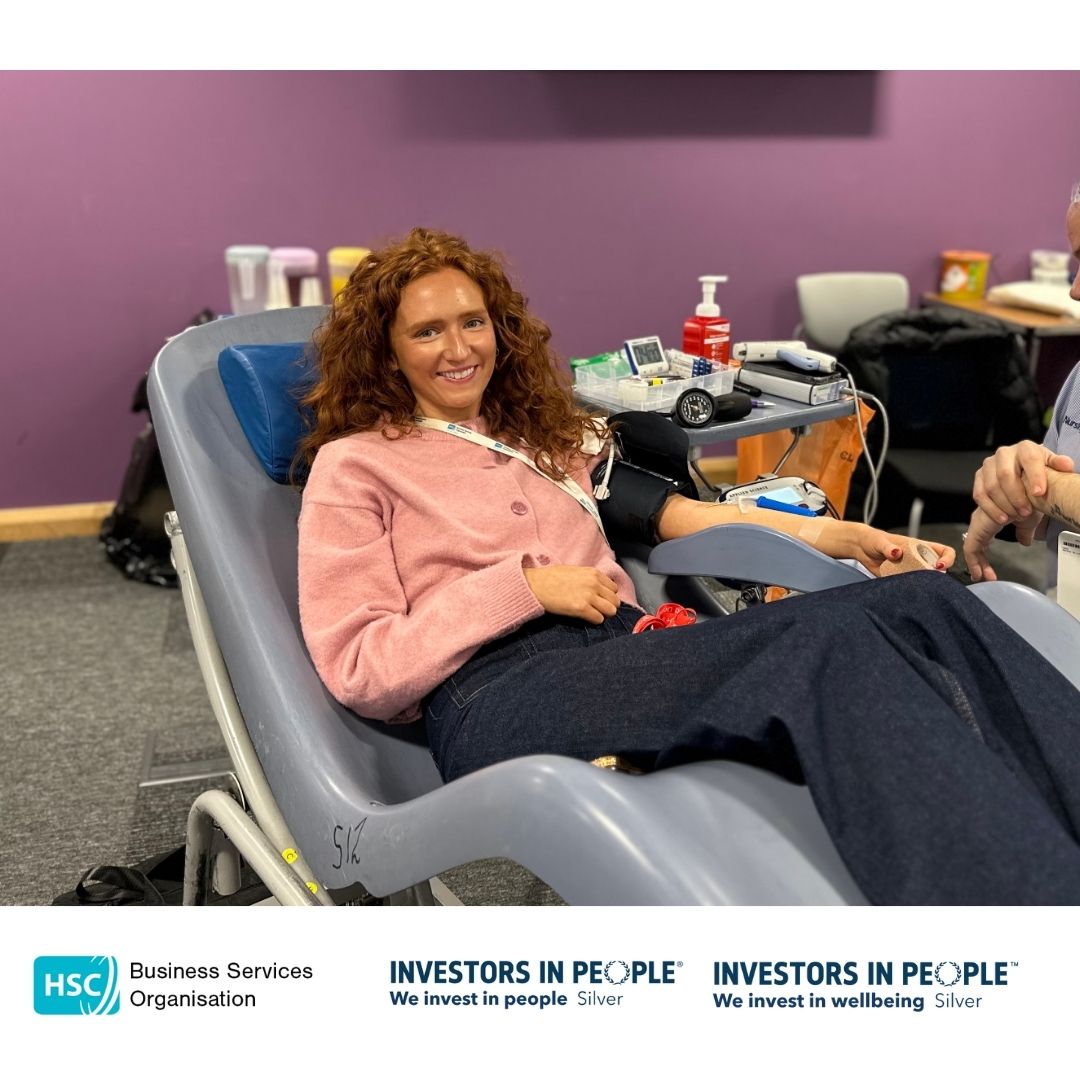 BSO Colleagues leading by example and are reclining on a chair, donating blood. They are holding a blue stress ball and appear relaxed, smiling at the camera. Medical equipment is visible nearby. The bottom of the image features logos for the Business Services Organisation and Investors in People, along with the text "We invest in people Silver" and "We invest in wellbeing Silver.