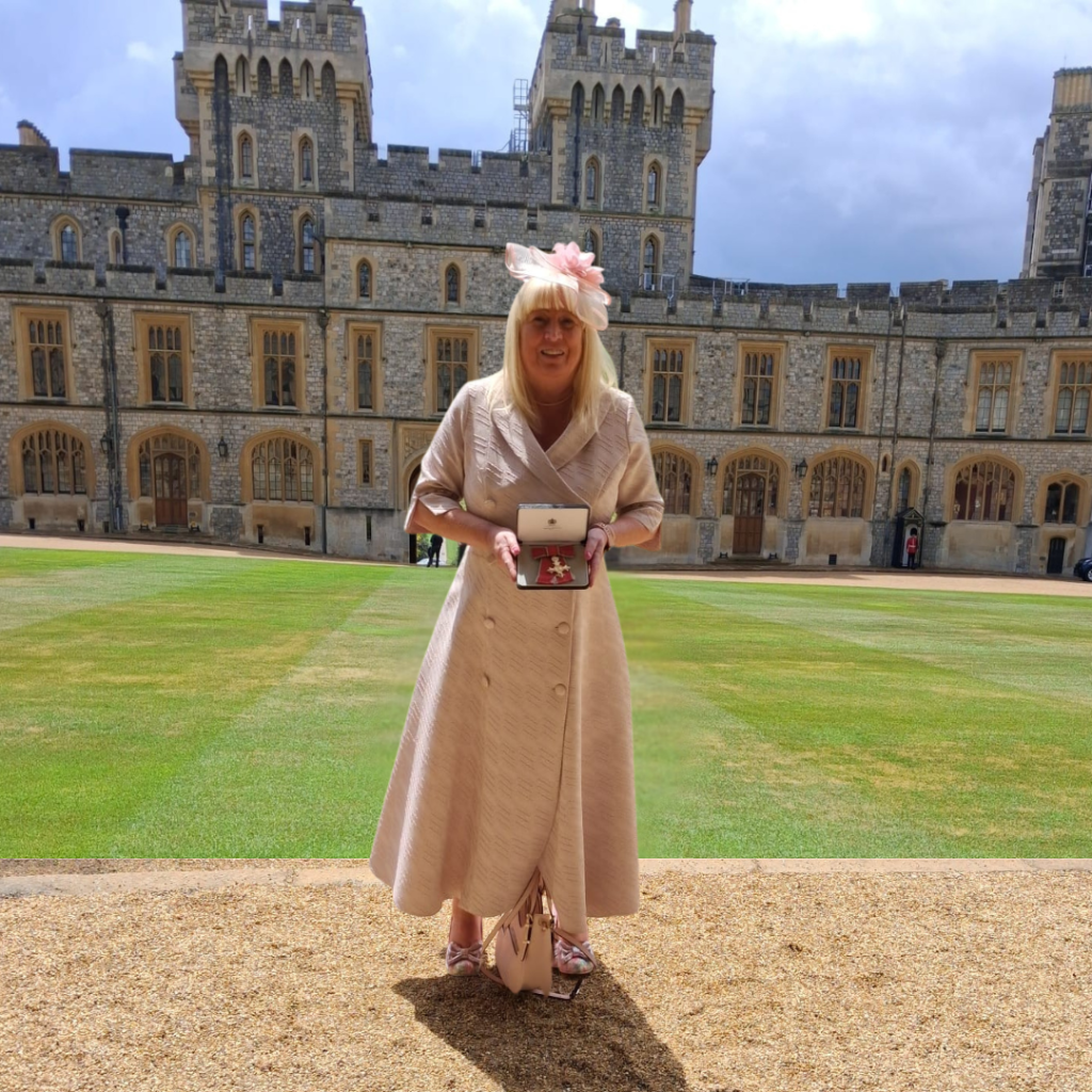 Women in pink dress and hat, standing in front of Windsor Castle with green grass and a gravel path. Women is hold an MBE award