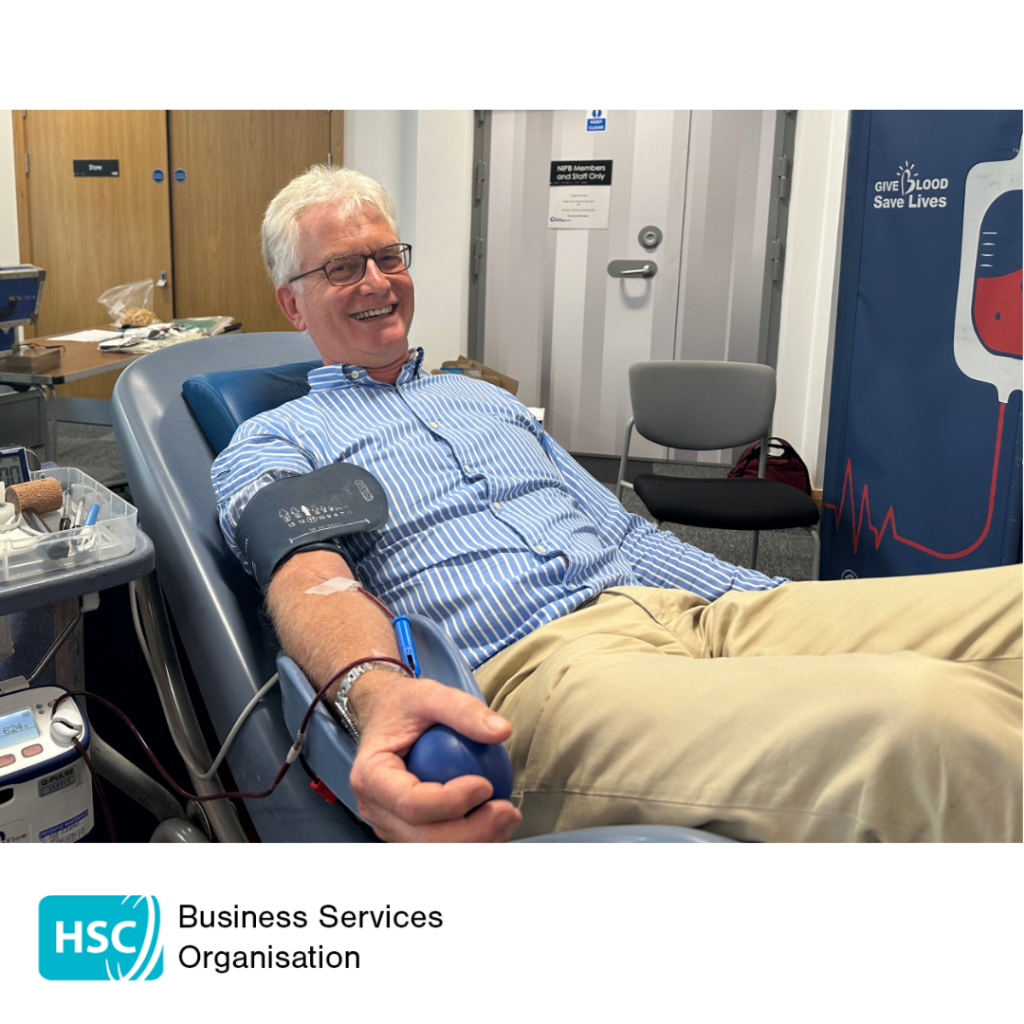 Person donating blood in a donor chair while holding a stress ball during a blood donation session. Donation equipment is connected to one arm. A "Give Blood Save Lives" display can be seen in the background. HSC Business Services Organisation logo displayed below the photograph.