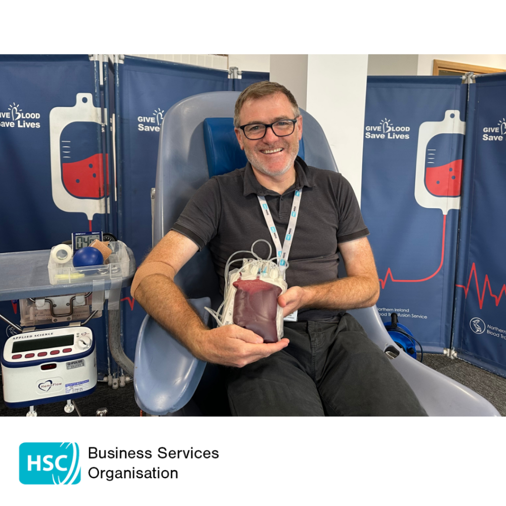 Blood donor seated in a donation chair holding a filled blood collection bag after donating blood. "Give Blood Save Lives" displays and donation equipment are visible in the background. HSC Business Services Organisation logo displayed below the photograph.