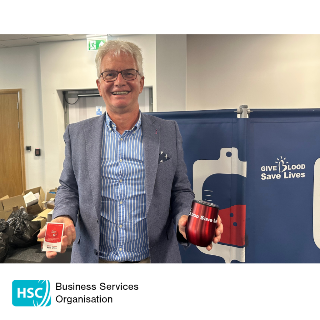 Person standing at a blood donation venue holding blood donor promotional items in front of a "Give Blood Save Lives" display. HSC Business Services Organisation logo displayed below the photograph.