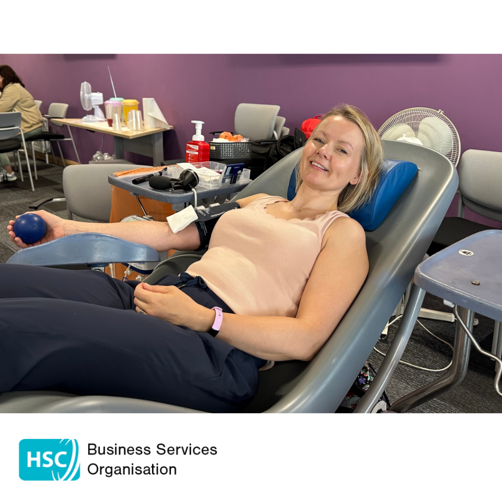 Blood donor reclining in a donation chair during a blood donation session, holding a stress ball while donation equipment is attached to one arm. A refreshment area and seating are visible in the background. HSC Business Services Organisation logo displayed below the photograph.