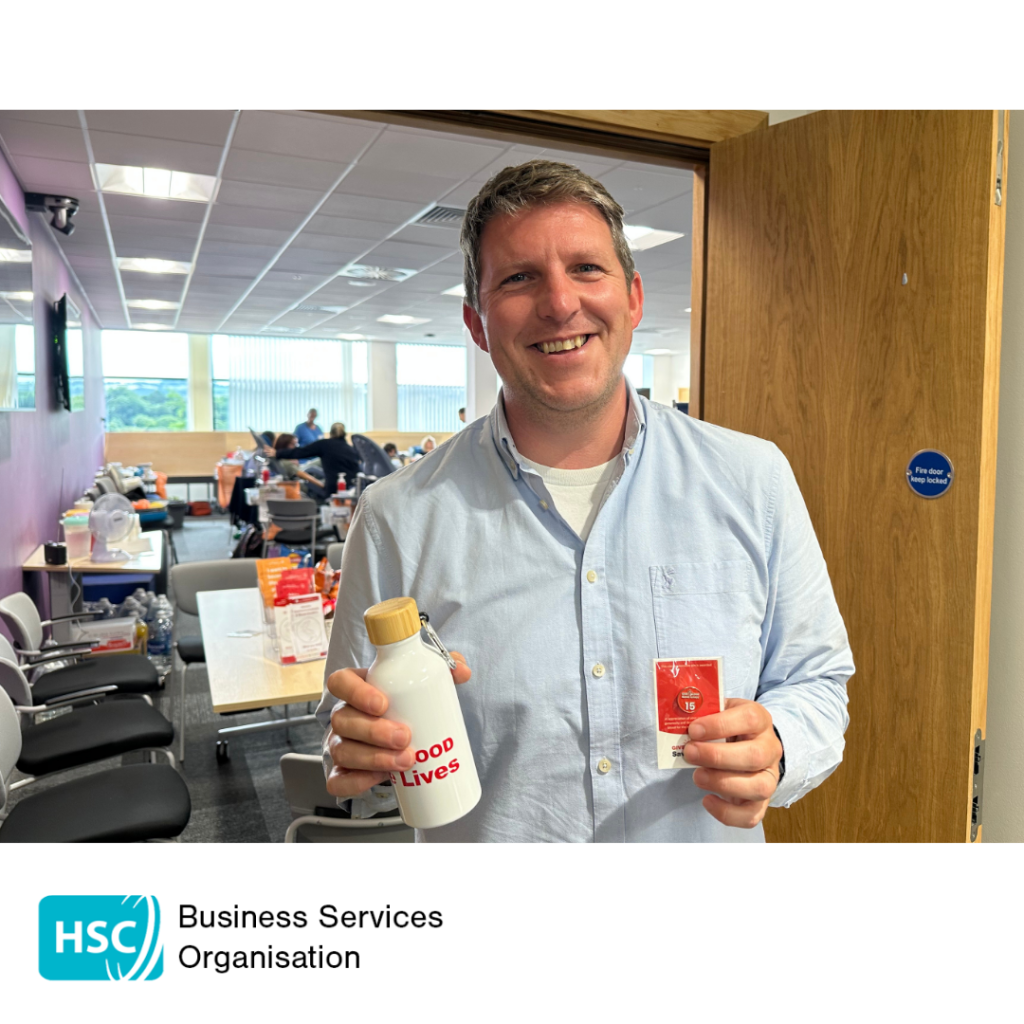 Staff member standing at a blood donation venue holding a reusable water bottle and a blood donor recognition badge. Blood donation chairs and facilities are visible in the background. HSC Business Services Organisation logo displayed below the photograph.