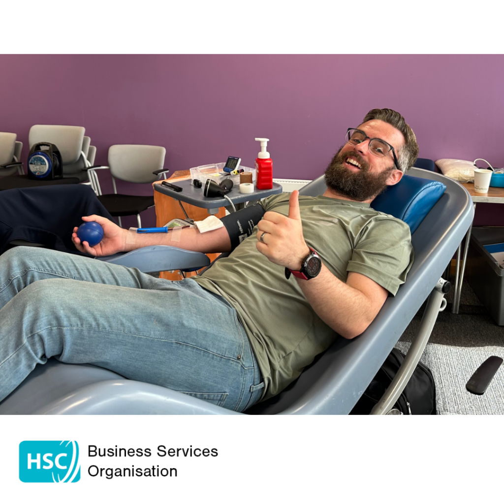 Blood donor reclining in a donation chair during a blood donation session, holding a stress ball while donation equipment is attached to one arm. The donor is giving a thumbs-up gesture. HSC Business Services Organisation logo displayed below the photograph.