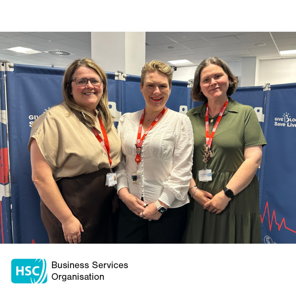 Three staff members standing together at a blood donation venue in front of "Give Blood Save Lives" displays. All are wearing staff identification lanyards. HSC Business Services Organisation logo displayed below the photograph.