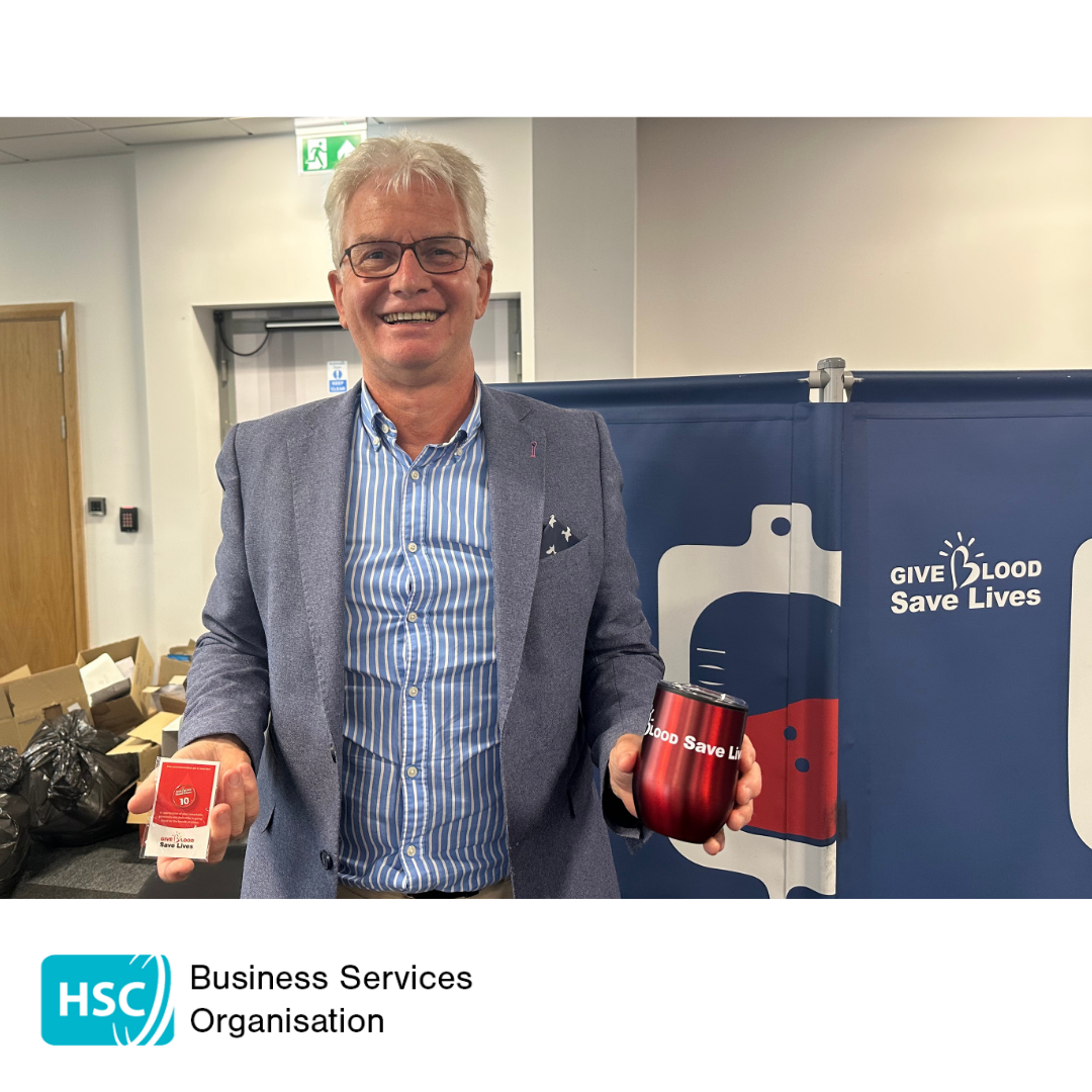 Person wearing a blue blazer and striped shirt stands at a blood donation awareness event, holding a red “Blood Save Lives” cup and a small promotional item. A blue backdrop behind the person features a blood bag graphic and the text “Give Blood Save Lives.” Cardboard boxes are visible in the background, and the HSC Business Services Organisation logo appears at the bottom of the image.