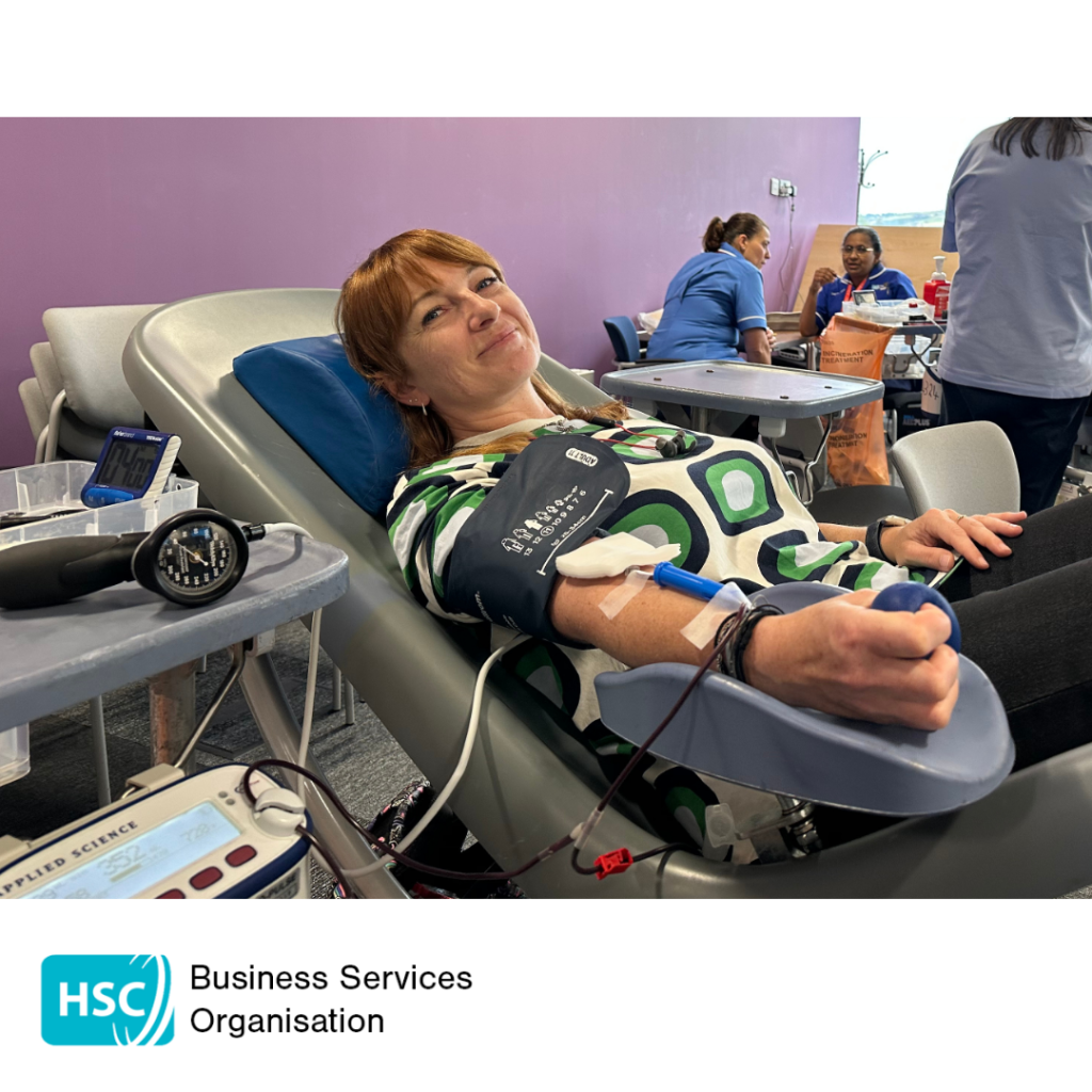 Person donating blood while reclining in a donor chair at a blood donation session. A blood pressure cuff and donation equipment are attached, with healthcare staff visible in the background. HSC Business Services Organisation logo displayed below the photograph.