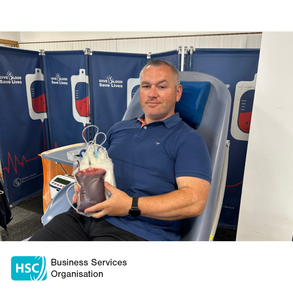 Blood donor seated in a donation chair holding a filled blood collection bag after donating blood. "Give Blood Save Lives" displays are visible in the background. HSC Business Services Organisation logo displayed below the photograph.