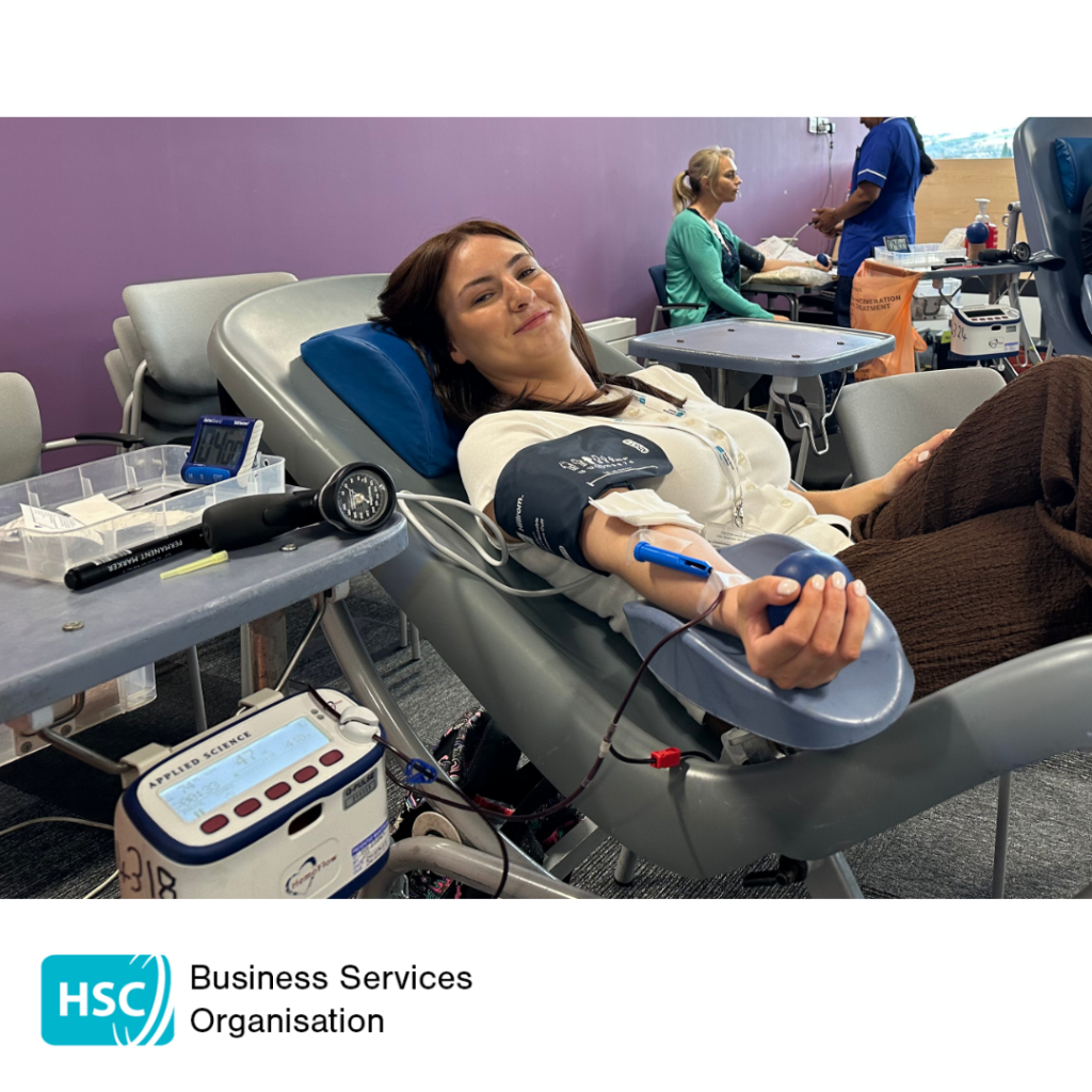 Blood donor donating blood while seated in a reclining chair and holding a stress ball. Blood donation equipment is attached to one arm, with healthcare staff and another donor visible in the background. HSC Business Services Organisation logo displayed below the photograph.