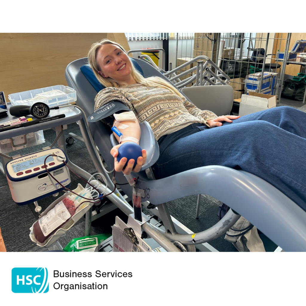 Blood donor reclining in a donation chair while giving blood and holding a stress ball. Donation equipment and a collection bag are visible beside the chair. HSC Business Services Organisation logo displayed below the photograph.