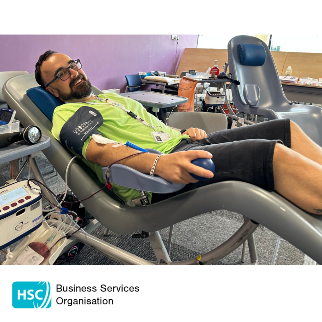 Blood donor reclining in a donation chair while giving blood and holding a stress ball. A blood collection bag and donation equipment are visible beside the chair. HSC Business Services Organisation logo displayed below the photograph.