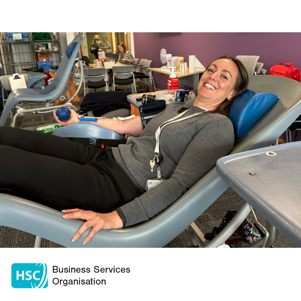 Blood donor seated in a reclining donation chair during a blood donation session, holding a stress ball with donation equipment attached to one arm. Tables, chairs and donation facilities are visible in the background. HSC Business Services Organisation logo displayed below the photograph.