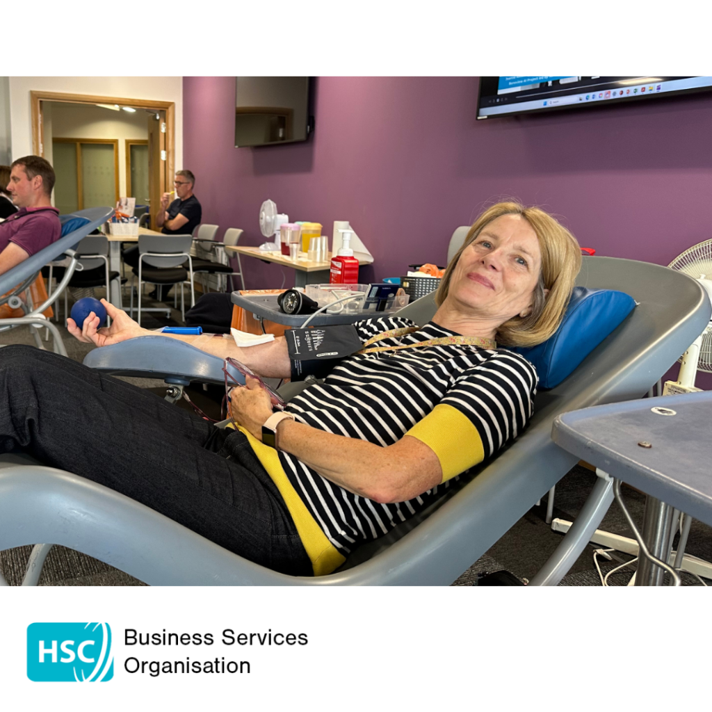 Blood donor reclining in a donation chair during a blood donation session, holding a stress ball while donation equipment is attached to one arm. Other donors and a refreshment area are visible in the background. HSC Business Services Organisation logo displayed below the photograph.