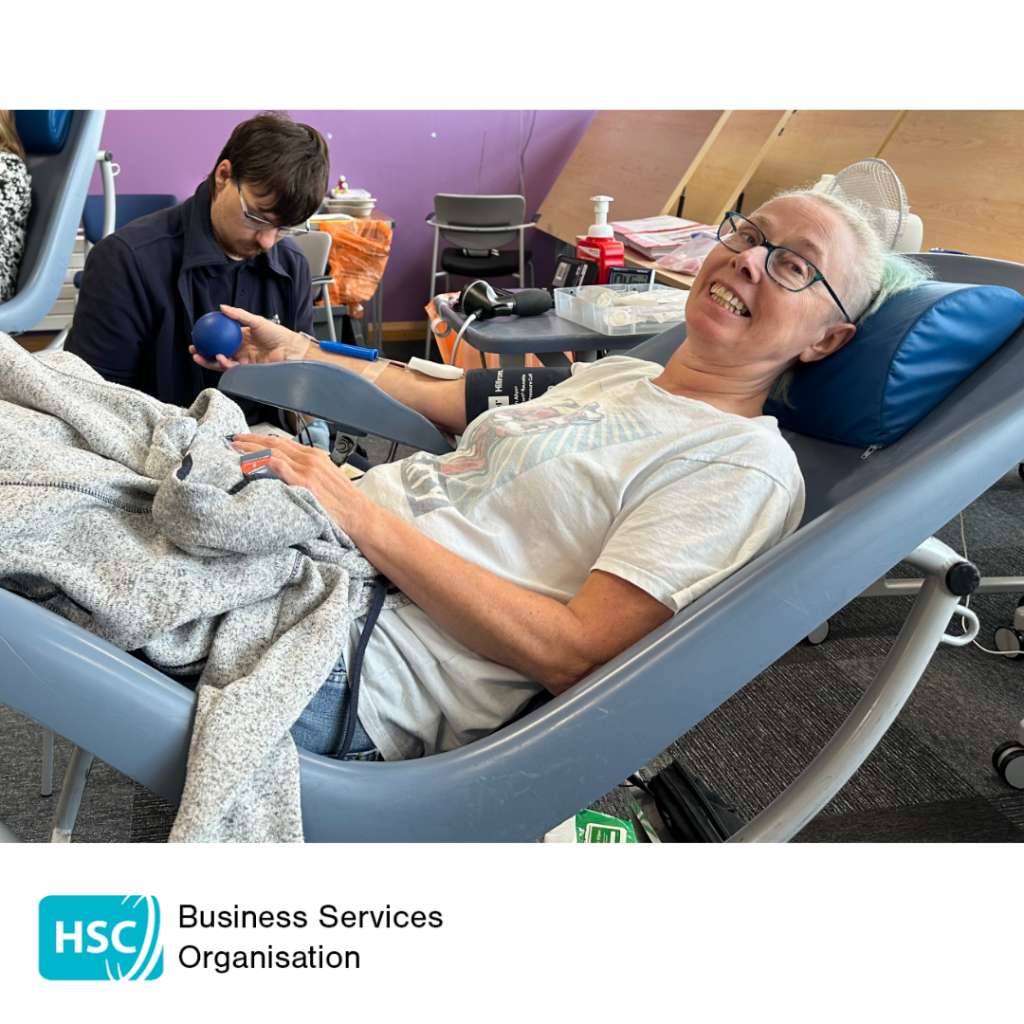 Blood donor reclining in a donation chair during a blood donation session, holding a stress ball while donation equipment is attached to one arm. Another blood donor and donation equipment are visible in the background. HSC Business Services Organisation logo displayed below the photograph.