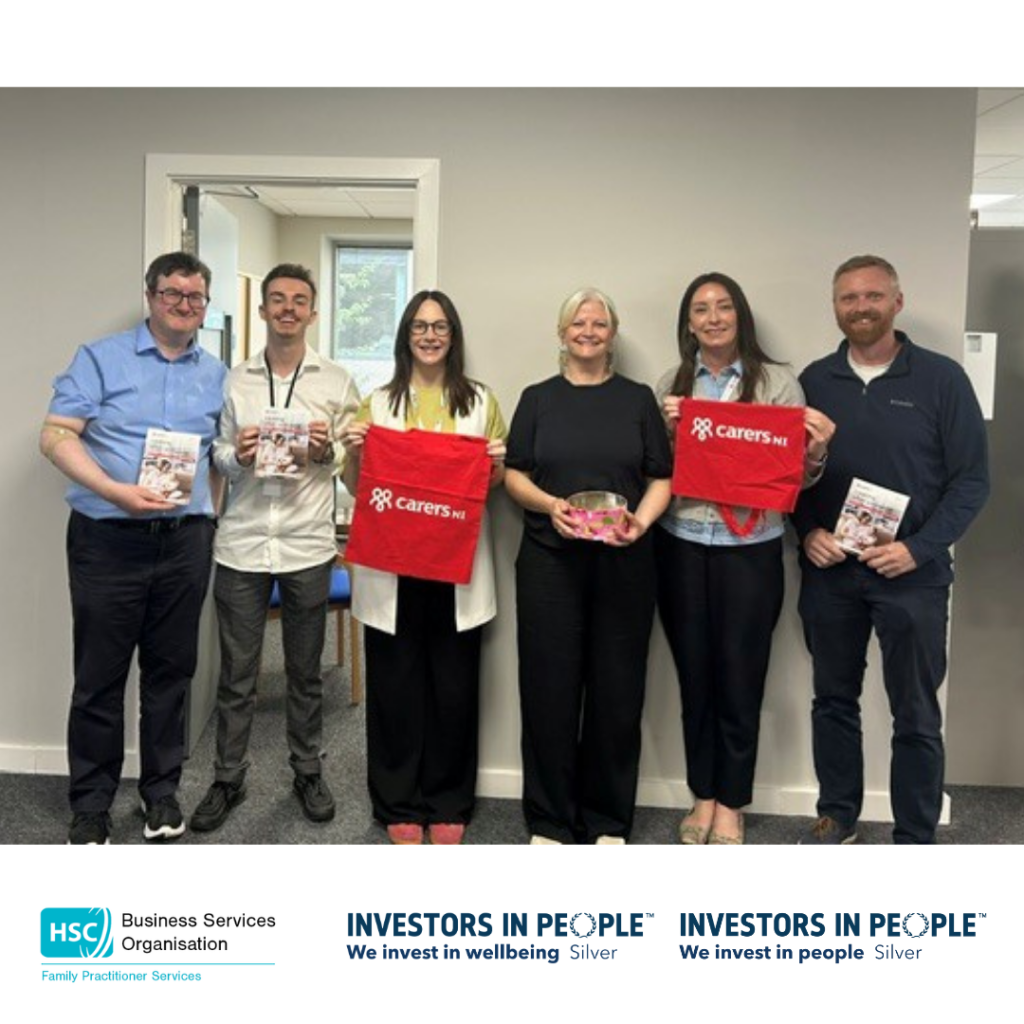 A group of six people stand together in an office, holding Carers NI information leaflets and red Carers NI tote bags. One person holds a bowl of promotional items. HSC Business Services Organisation and Investors in People Silver logos are displayed along the bottom of the image.