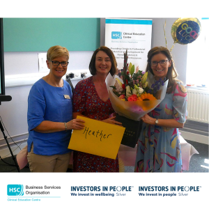 Three colleagues standing together at a Clinical Education Centre retirement celebration, presenting flowers and a card. The group is positioned in front of a Clinical Education Centre banner, with a bouquet of flowers, balloons and retirement gifts visible in the room.