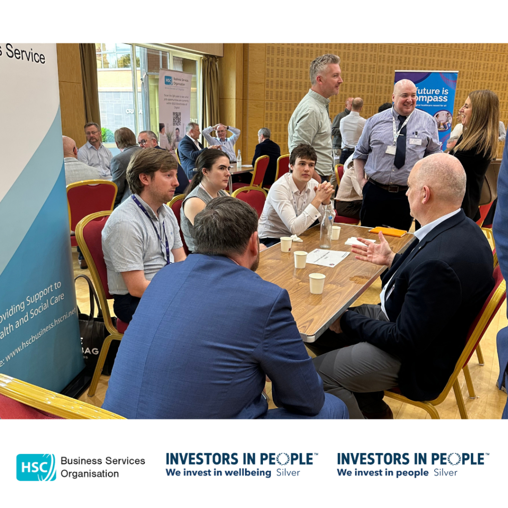 Attendees take part in a small-group networking discussion at the Public Procurement – Meet the Buyer Event. Several participants are seated around a table in a conference venue, discussing opportunities and sharing information, while other attendees network at nearby tables in the background. Information sheets, cups and water bottles are visible on the table. Business Services Organisation and partner organisation banners are displayed around the room. The HSC Business Services Organisation and Investors in People Silver logos appear along the bottom of the image.