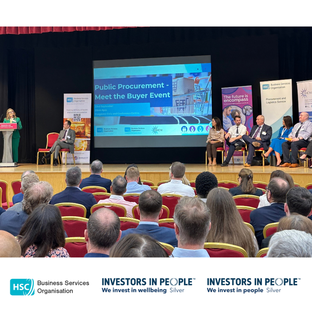 Audience members sit in a conference hall facing a stage at the Public Procurement – Meet the Buyer Event. A speaker stands at a lectern on the left of the stage, while a panel of attendees is seated on the right. A large screen behind them displays the event title and details. Promotional banners from partner organisations are positioned around the stage. The HSC Business Services Organisation and Investors in People Silver logos are displayed along the bottom of the image.