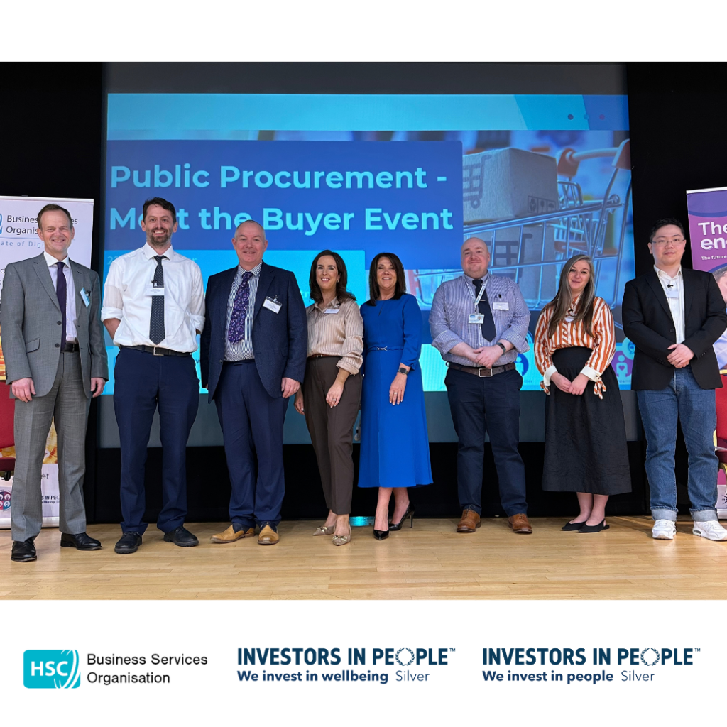 Eight people stand on a stage in front of a large presentation screen displaying the text “Public Procurement – Meet the Buyer Event.” The group is dressed in business attire and poses for a photograph at the event. Banners are visible on either side of the stage, and the HSC Business Services Organisation and Investors in People Silver logos appear along the bottom of the image.