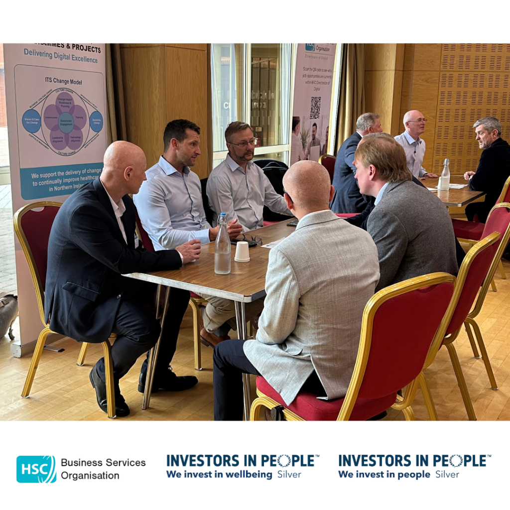 Attendees take part in a networking discussion at the Public Procurement – Meet the Buyer Event. Six participants are seated around a table in a conference venue, engaged in conversation, with water bottles and notepads on the table. Additional small-group discussions are taking place at tables in the background. A Business Services Organisation Digital Directorate banner is visible behind the group. The HSC Business Services Organisation and Investors in People Silver logos appear along the bottom of the image.