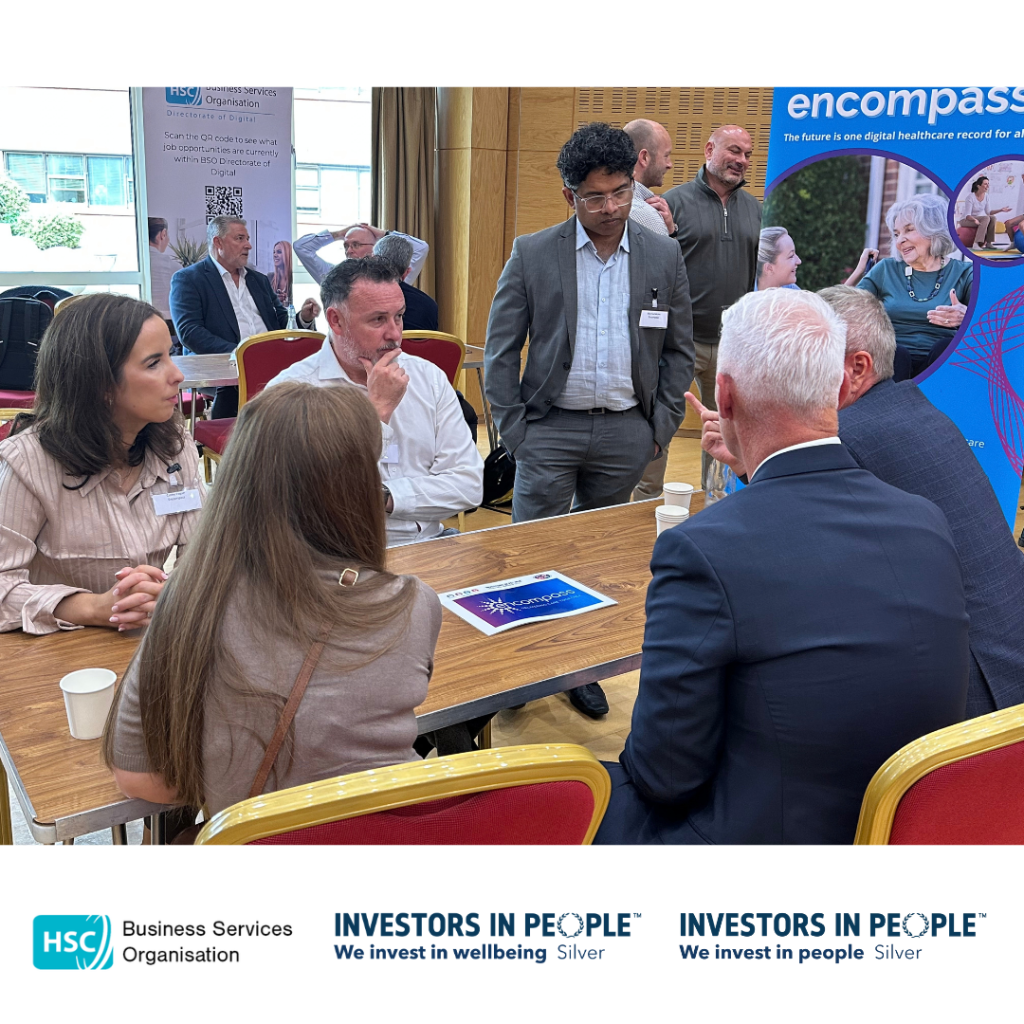 Attendees participate in a round-table networking discussion during the Public Procurement – Meet the Buyer Event. Several participants are seated around a table while others stand nearby engaging in conversation. Event information sheets and refreshments are visible on the table. Banners promoting the HSC Business Services Organisation Digital Directorate and the Encompass digital healthcare programme are displayed in the room. Additional networking discussions are taking place in the background. The HSC Business Services Organisation and Investors in People Silver logos appear along the bottom of the image.