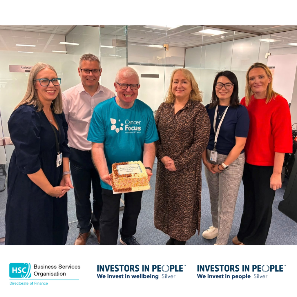A group of six colleagues stand together in an office meeting room with glass walls. The person in the centre is holding a decorated celebration cake and wearing a turquoise Cancer Focus Northern Ireland T-shirt. The group is gathered for a presentation or retirement celebration. At the bottom of the graphic are the logos for the Health and Social Care Business Services Organisation Directorate of Finance and Investors in People Silver accreditation. Visible text on the cake reads: “Congratulations on your retirement Peter.”