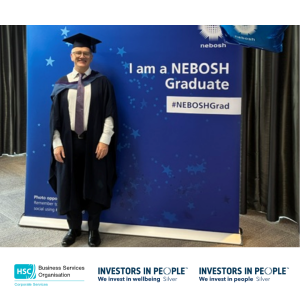 Person in graduation robes standing beside a NEBOSH Graduate display board at a diploma graduation ceremony.