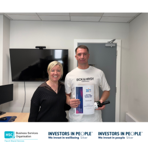 Two colleagues standing in an office holding a National Payroll Week 2026 Payroll Dedication Award certificate and a small presentation bottle. A wall-mounted screen and office door are visible in the background.