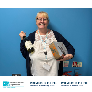 Colleague standing against a blue wall holding a bottle of wine and a gift tin. The image is branded with Business Services Organisation Payroll Shared Services and Investors in People Silver logos along the bottom
